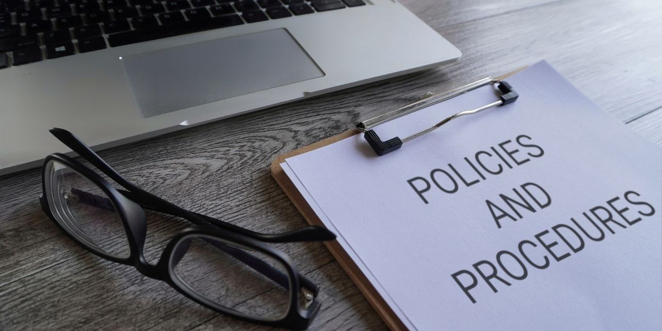 Paper clipboard with text POLICIES AND PROCEDURES on table with laptop and glasses.